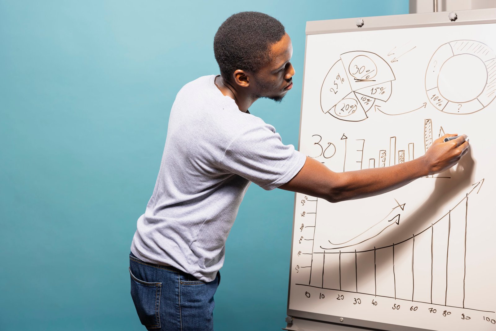 African american male professional in casual clothing drawing graphs and charts on a sketch board. Black man standing next to whiteboard and preparing data for presentation in blue background studio.